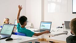 Junge sitzt in einem Klassenzimmer vor einem Laptop und hebt die Hand