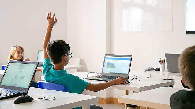 Junge sitzt in einem Klassenzimmer vor einem Laptop und hebt die Hand