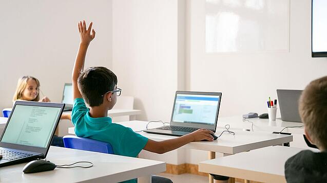 Junge sitzt in einem Klassenzimmer vor einem Laptop und hebt die Hand