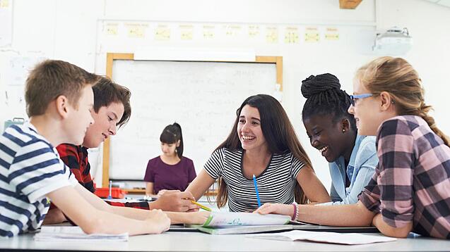 Gut gelaunte Kinder sitzen in der Schule um einen Tisch und lernen.
