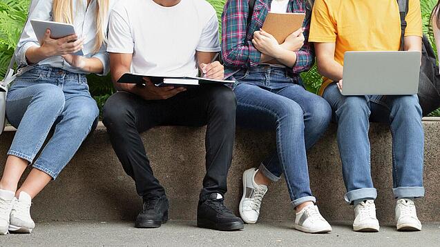 Mehrere Jugendliche sitzen nebeneinander auf einer Mauer und schauen in ihre Laptops, Tablets oder Hefte.