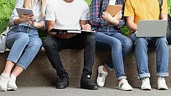 Mehrere Jugendliche sitzen nebeneinander auf einer Mauer und schauen in ihre Laptops, Tablets oder Hefte.