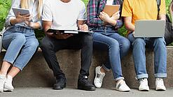 Mehrere Jugendliche sitzen nebeneinander auf einer Mauer und schauen in ihre Laptops, Tablets oder Hefte.