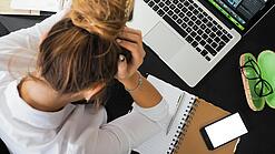 Eine junge Frau stützt frustriert ihren Kopf in ihre Hände, vor ihr auf dem Tisch ein Laptop und ein Notizbuch.