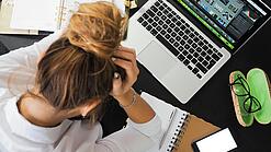 Eine junge Frau stützt frustriert ihren Kopf in ihre Hände, vor ihr auf dem Tisch ein Laptop und ein Notizbuch.