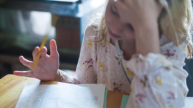 Ein junges Mädchen sitzt an einem Tisch und blickt frustriert auf ein Heft, das vor ihr liegt; ihren Kopf stützt sie mit einer Hand.
