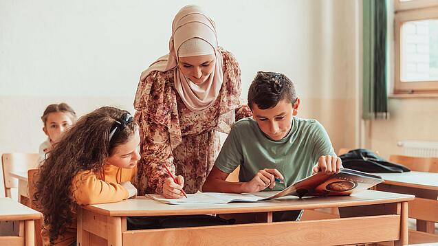 Eine Lehrerin beugt sich über zwei Schülerinnen und Schüler, die an etwas arbeiten.