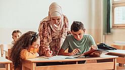 Eine Lehrerin beugt sich über zwei Schülerinnen und Schüler, die an etwas arbeiten.