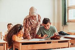 Eine Lehrerin beugt sich über zwei Schülerinnen und Schüler, die an etwas arbeiten.