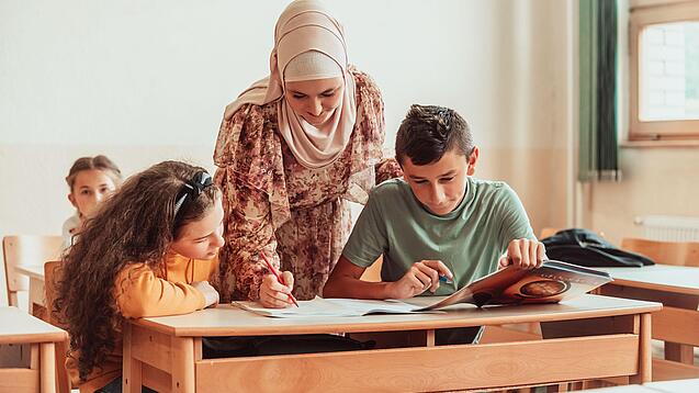 Eine Lehrerin beugt sich über zwei Schülerinnen und Schüler, die an etwas arbeiten.