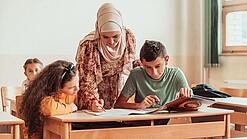 Eine Lehrerin beugt sich über zwei Schülerinnen und Schüler, die an etwas arbeiten.