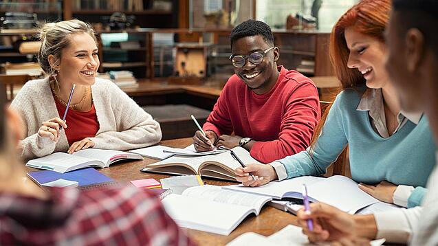 Mehrere Studierende sitzen an einem Tisch und arbeiten gut gelaunt in ihren Unterlagen.