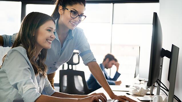 Zwei junge Frauen schauen lächelnd auf den Bildschirm eines Computers.