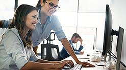 Zwei junge Frauen schauen lächelnd auf den Bildschirm eines Computers.