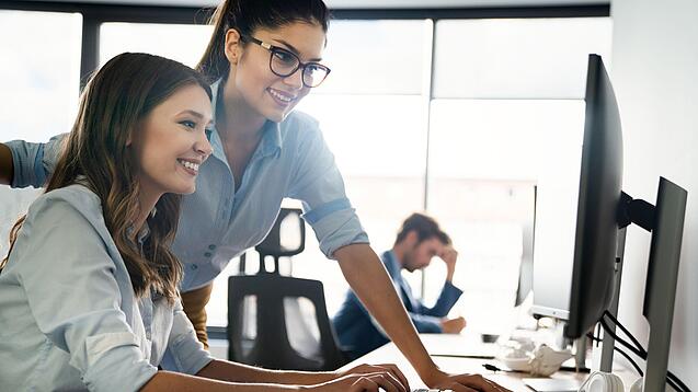 Zwei junge Frauen schauen lächelnd auf den Bildschirm eines Computers.