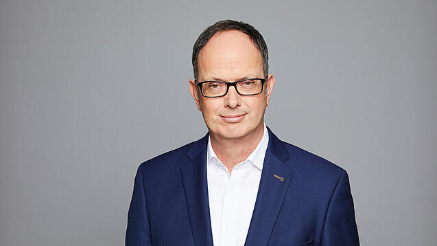 Frank Thalhofer lächelt vor einem grauen Hintergrund in die Kamera. 