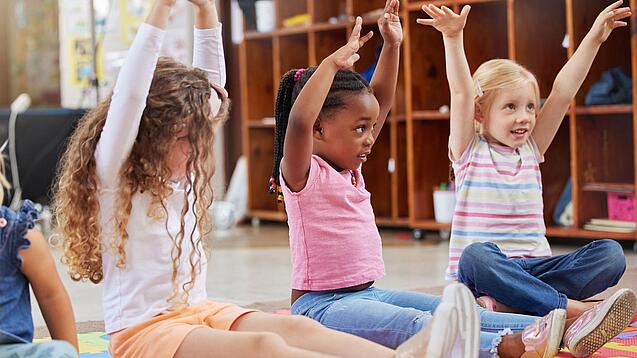 Mehrere Kita-Kinder sitzen auf dem Boden und strecken ihre Arme in die Höhe.