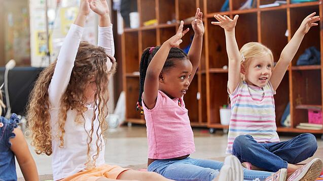 Mehrere Kita-Kinder sitzen auf dem Boden und strecken ihre Arme in die Höhe.