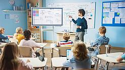Ein Lehrer steht an einem Bildschirm und erklärt den an ihren Tischen sitzenden Schülerinnen und Schülern etwas.