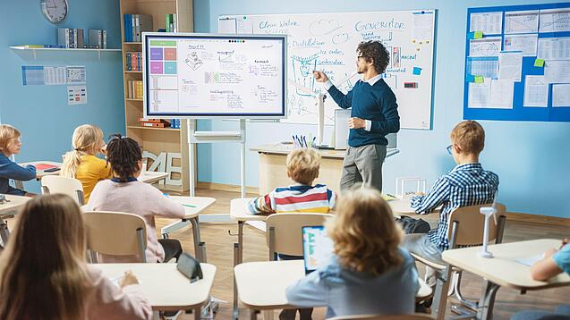 Ein Lehrer steht an einem Bildschirm und erklärt den an ihren Tischen sitzenden Schülerinnen und Schülern etwas.