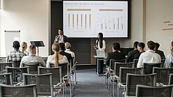 Mehrere Jugendliche und junge Erwachsene sitzen in einem Raum auf Stühlen und verfolgen eine Präsentation.