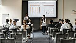 Mehrere Jugendliche und junge Erwachsene sitzen in einem Raum auf Stühlen und verfolgen eine Präsentation.