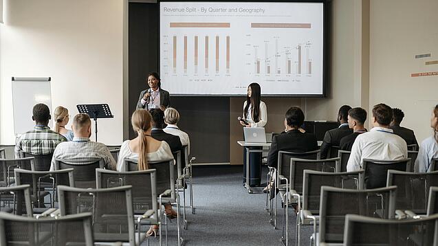 Mehrere Jugendliche und junge Erwachsene sitzen in einem Raum auf Stühlen und verfolgen eine Präsentation.