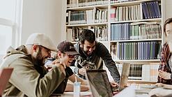 Gruppe junger Menschen sitzt lachen vor Laptops und einem Bücherregal.
