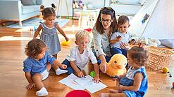 Eine Erzieherin sitzt mit Kindern auf dem Boden und malt.