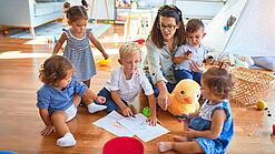 Eine Erzieherin sitzt mit Kindern auf dem Boden und malt.