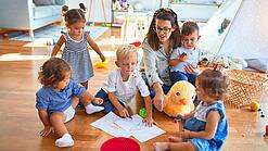 Eine Erzieherin sitzt mit Kindern auf dem Boden und malt.