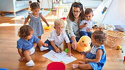 Eine Erzieherin sitzt mit Kindern auf dem Boden und malt.