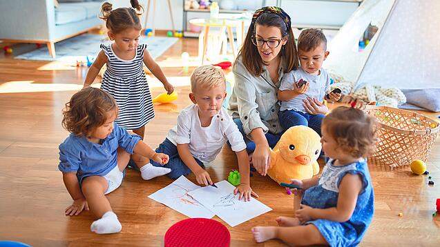 Eine Erzieherin sitzt mit Kindern auf dem Boden und malt.