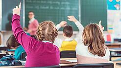 Blick von hinten auf eine Schulklasse, in der sich mehrere Kinder melden; eine Lehrerin steht an der Tafel.