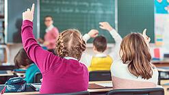 Blick von hinten auf eine Schulklasse, in der sich mehrere Kinder melden; eine Lehrerin steht an der Tafel.