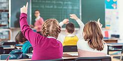 Blick von hinten auf eine Schulklasse, in der sich mehrere Kinder melden; eine Lehrerin steht an der Tafel.