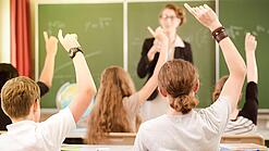 Jugendliche sitzen in einer Schulklasse und melden sich, im Hintergrund steht eine Lehrerin an der Tafel.