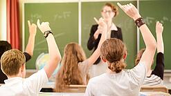 Jugendliche sitzen in einer Schulklasse und melden sich, im Hintergrund steht eine Lehrerin an der Tafel.