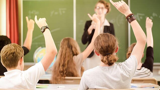 Jugendliche sitzen in einer Schulklasse und melden sich, im Hintergrund steht eine Lehrerin an der Tafel.