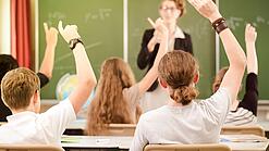 Jugendliche sitzen in einer Schulklasse und melden sich, im Hintergrund steht eine Lehrerin an der Tafel.