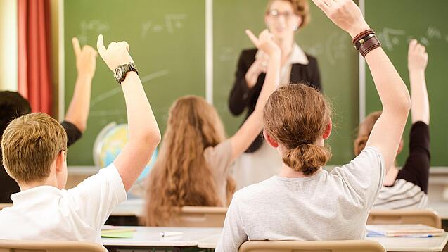 Jugendliche sitzen in einer Schulklasse und melden sich, im Hintergrund steht eine Lehrerin an der Tafel.