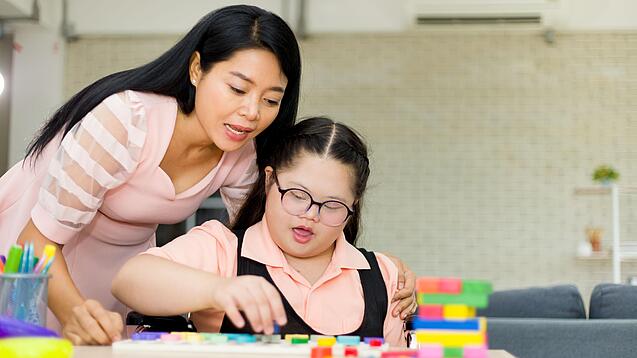Mädchen mit Downsyndrom sitzt an einem Tisch und spielt mit Bauklötzen, junge Frau beugt sich über sie