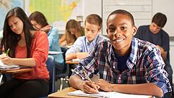 Mehrere Schülerinnen und Schüler sitzen an Tischen in einer Klasse und arbeiten in ihren Heften; vorne lächelt ein Schüler in die Kamera.