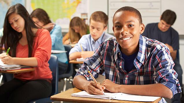 Mehrere Schülerinnen und Schüler sitzen an Tischen in einer Klasse und arbeiten in ihren Heften; vorne lächelt ein Schüler in die Kamera.
