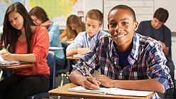 Mehrere Schülerinnen und Schüler sitzen an Tischen in einer Klasse und arbeiten in ihren Heften; vorne lächelt ein Schüler in die Kamera.