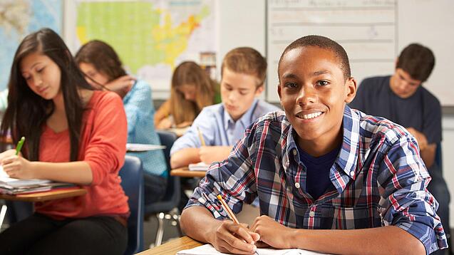 Mehrere Schülerinnen und Schüler sitzen an Tischen in einer Klasse und arbeiten in ihren Heften; vorne lächelt ein Schüler in die Kamera.