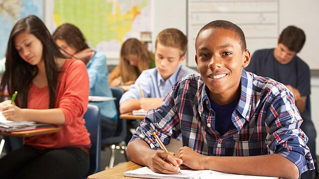 Mehrere Schülerinnen und Schüler sitzen an Tischen in einer Klasse und arbeiten in ihren Heften; vorne lächelt ein Schüler in die Kamera.