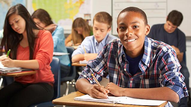 Mehrere Schülerinnen und Schüler sitzen an Tischen in einer Klasse und arbeiten in ihren Heften; vorne lächelt ein Schüler in die Kamera.