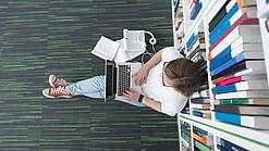 Junger Mensch sitzt mit einem Laptop auf dem Schoß vor einem Bücherregal auf dem Boden.