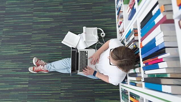 Junger Mensch sitzt mit einem Laptop auf dem Schoß vor einem Bücherregal auf dem Boden.